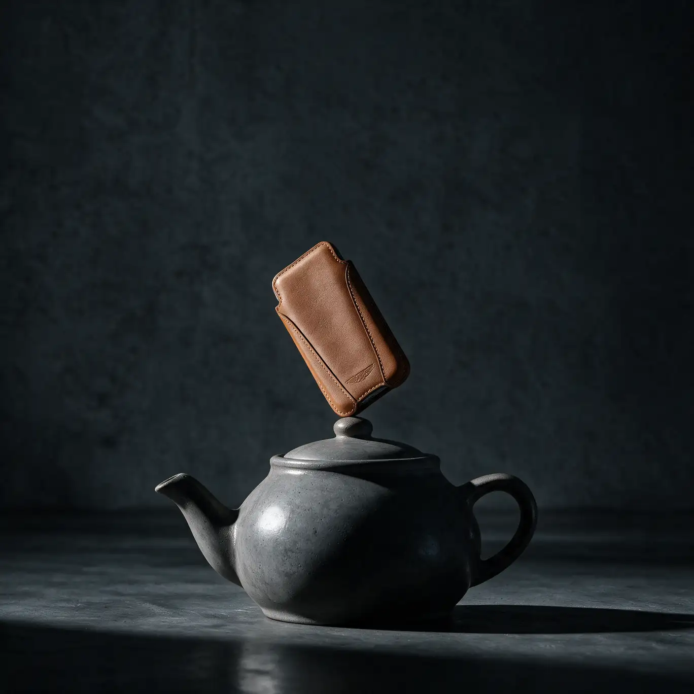 A tan leather phone sleeve balanced upright on the lid of a grey stone teapot